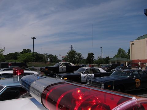 1991 Ford Crown Victoria - A collection of police vehicles, including a 1979 Ford LTD, are visible in a parking lot on a partly cloudy day.