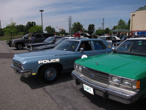 1991 Ford Crown Victoria - A lineup of police vehicles, a blue sedan and a green sedan, parked outdoors on a sunny day.