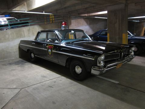 1991 Ford LTD Crown Victoria - black Pontiac police sedan with a door shield emblem and red roof beacon sits in a dim concrete parking garage.