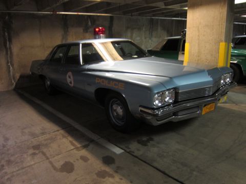 1991 Ford LTD Crown Victoria - light blue 1970s Buick police sedan with 'POLICE' lettering and a red roof beacon, parked in a concrete garage.
