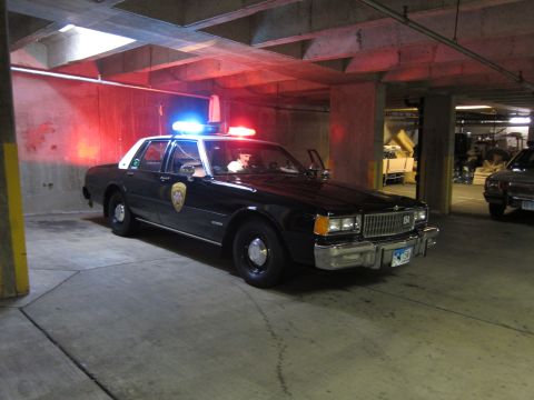 1991 Ford LTD Crown Victoria - dark Chevrolet Caprice police sedan with illuminated red roof lights and a door shield, parked in a garage.