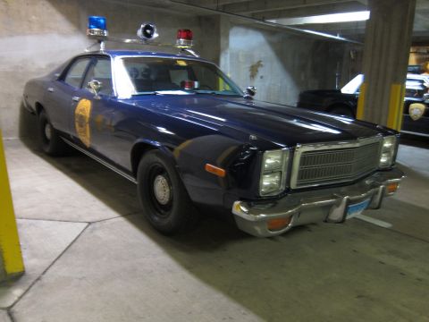 1991 Ford LTD Crown Victoria - dark blue 1970s Plymouth police sedan with a gold door shield and roof beacons, parked in a concrete garage.