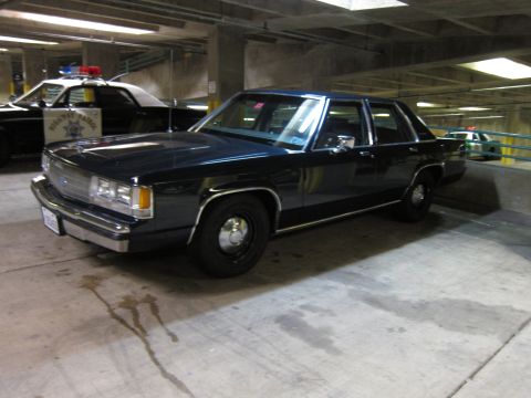1991 Ford LTD Crown Victoria - dark Ford LTD Crown Victoria police sedan with a red roof beacon parked indoors beside a black-and-white patrol car.