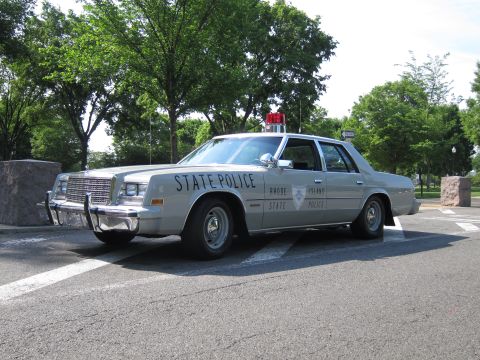 1991 Ford LTD Crown Victoria - silver Chrysler-style 'Rhode Island State Police' sedan with a single red roof beacon, parked outdoors among trees.