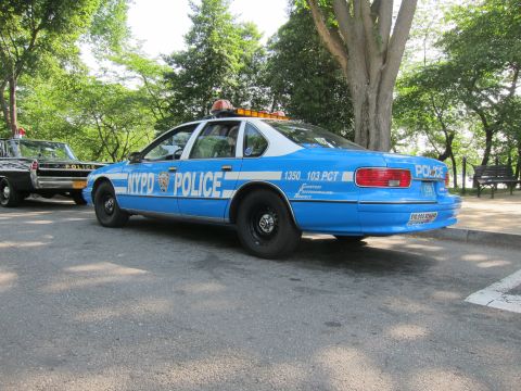 1991 Ford LTD Crown Victoria - blue-and-white Chevrolet Caprice marked 'NYPD POLICE' with roof lights, viewed from the rear on a tree-lined street.