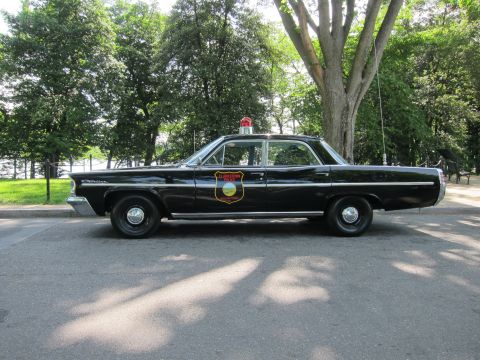 1991 Ford LTD Crown Victoria - black vintage Pontiac police sedan with a door shield and red roof beacon, shown in side profile on a shaded street.