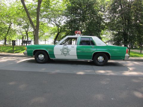1991 Ford LTD Crown Victoria - green-and-white Ford LTD sheriff sedan with a star emblem and red roof beacon, shown in side profile outdoors.