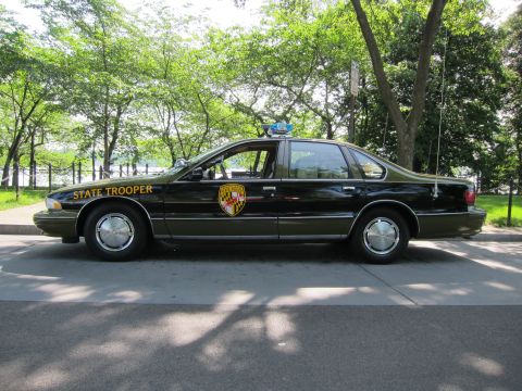 1991 Ford LTD Crown Victoria - black Chevrolet Caprice 'STATE TROOPER' sedan with a gold shield and blue roof light, parked in side profile outdoors.