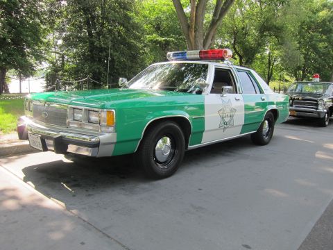 1991 Ford LTD Crown Victoria - green-and-white Ford LTD sheriff sedan with a star emblem and roof lights, parked outdoors beside an older patrol car.