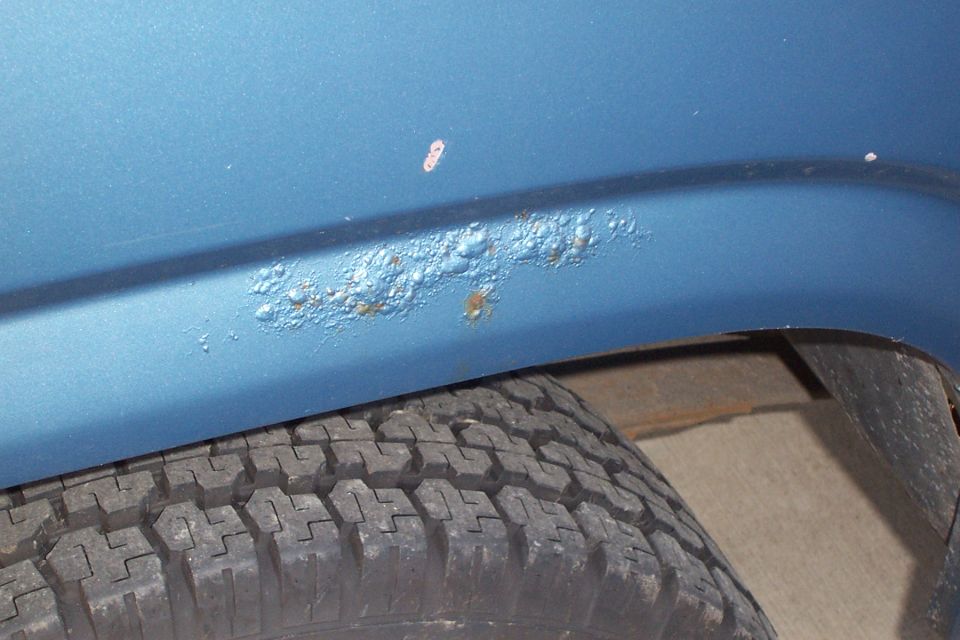 Close-up of rust bubbles forming on the lower fender of a blue vehicle, with a tire visible below