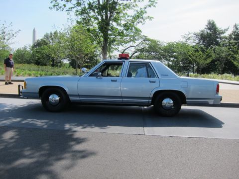 1991 Ford LTD Crown Victoria - silver Ford LTD Crown Victoria police sedan shown in side profile with a red roof beacon, parked near a tree-lined road with a monument beh…