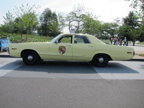 1991 Ford LTD Crown Victoria - pale yellow early-1970s Dodge police sedan bearing a state-police shield emblem and red roof beacon, shown in side profile.