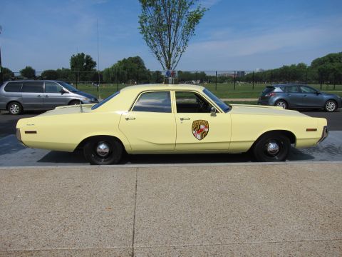 1991 Ford LTD Crown Victoria - pale yellow vintage Dodge police sedan with a state-police shield door emblem and roof antenna, parked along a grassy field.