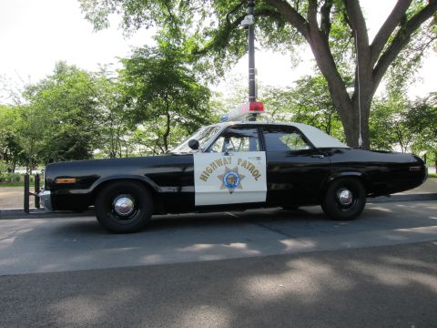 1991 Ford LTD Crown Victoria - black-and-white 1970s Dodge Highway Patrol sedan with a single red roof beacon, shown in side profile under trees.