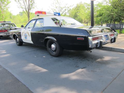 1991 Ford LTD Crown Victoria - black-and-white 1970s police sedan lettered POLICE with a red roof light bar, parked rear-quarter to camera in a row of cruisers.