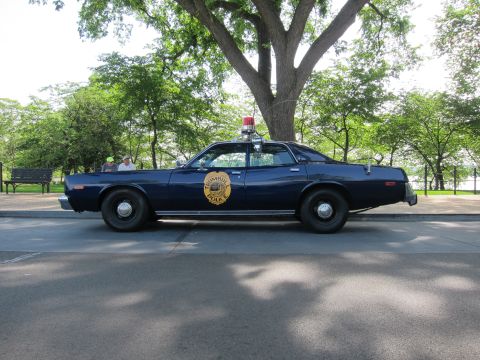 1991 Ford LTD Crown Victoria - dark blue 1970s police sedan with a gold circular highway-patrol emblem and red roof beacon, shown in side profile under a large tree.