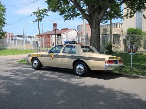 1991 Ford LTD Crown Victoria - tan State Police sedan with a state shield emblem and roof light bar, parked in front of fencing and industrial buildings.
