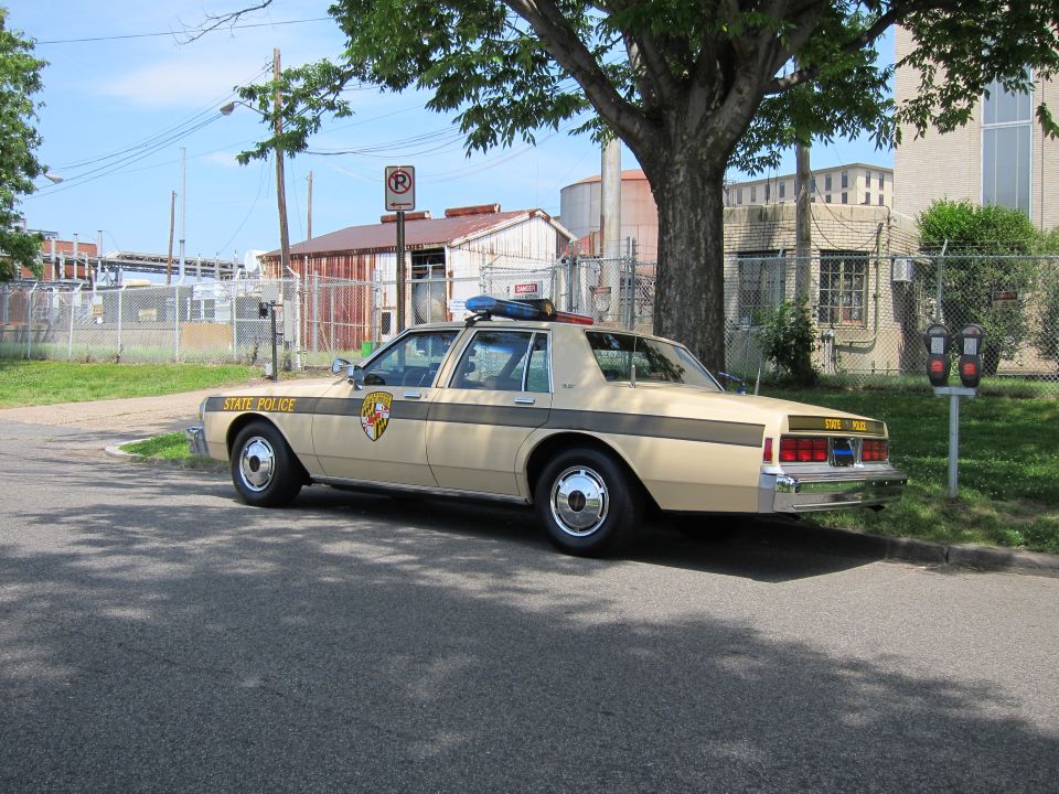 Tan State Police sedan with shield emblem, rear three-quarter, industrial backdrop