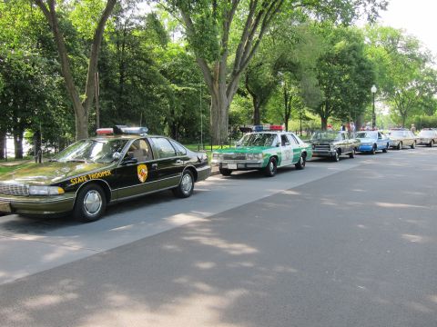 1991 Ford LTD Crown Victoria - row of police cruisers led by a black State Trooper Chevrolet Caprice with a shield emblem, lined along a tree-shaded road.