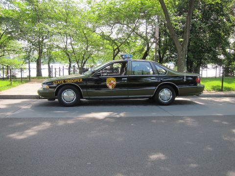 1991 Ford LTD Crown Victoria - black State Trooper Chevrolet Caprice sedan lettered State Trooper with a state shield emblem and roof light bar, shown in side profile.