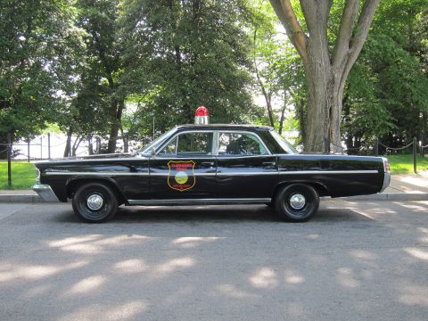 1991 Ford LTD Crown Victoria - black early-1960s Pontiac Catalina police sedan with a single red roof beacon and a state-police shield emblem, shown in side profile.