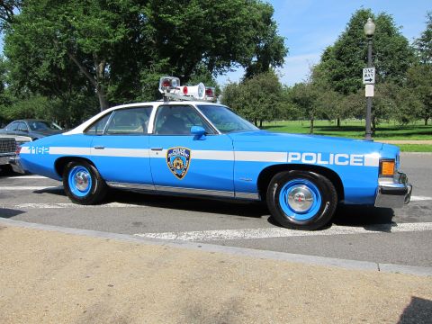 1991 Ford LTD Crown Victoria - blue-and-white 1970s Pontiac NYPD police sedan lettered POLICE with the NYPD shield, unit 1162 and roof light bar, parked at a curb.