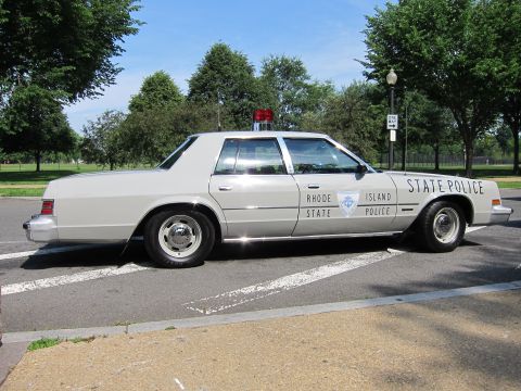 1991 Ford LTD Crown Victoria - Side profile of a silver Rhode Island State Police sedan with red roof beacon parked on a paved lot.