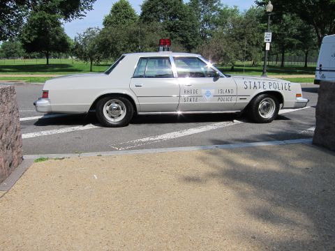 1991 Ford LTD Crown Victoria - silver Rhode Island State Police sedan with red roof light sits between low concrete barriers in a park.