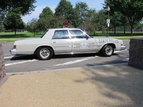 1991 Ford LTD Crown Victoria - Side view of a silver Rhode Island State Police four-door sedan parked near concrete posts in a park.