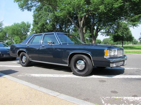 1991 Ford LTD Crown Victoria - Front-three-quarter view of a dark navy four-door sedan with plain hubcaps parked under trees in a lot.