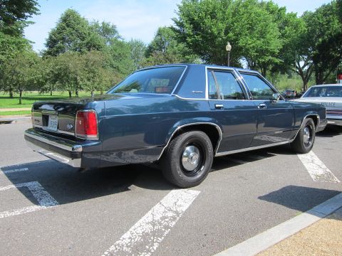 1991 Ford LTD Crown Victoria - Rear-three-quarter view of a dark blue four-door sedan in a lot, with a marked police car visible at right.