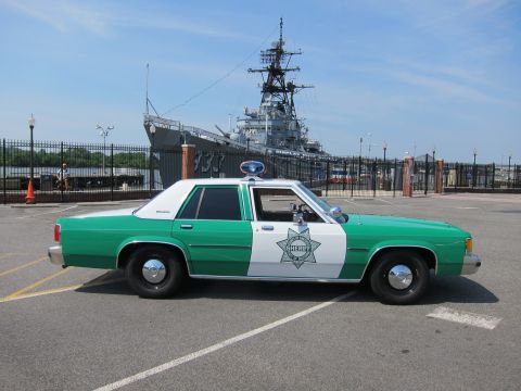1991 Ford LTD Crown Victoria - green-and-white Sheriff sedan with star decal sits in a lot in front of a moored gray naval warship.