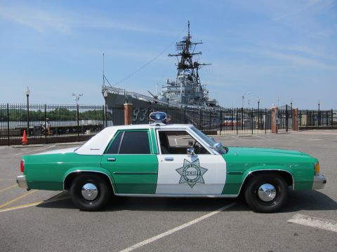 1991 Ford LTD Crown Victoria - Side profile of a green-and-white Sheriff sedan parked before a moored gray Navy warship marked 933.