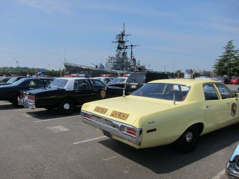 1991 Ford LTD Crown Victoria - pale-yellow State Police sedan and other police cars parked in a lot in front of a moored gray warship.