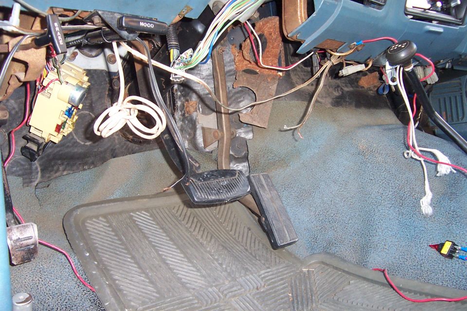 Interior shot of vehicle's dashboard wiring, pedals, and floor mat. Wires are exposed and appear rusty