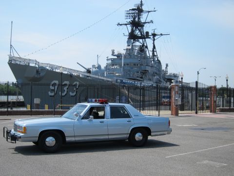 1991 Ford LTD Crown Victoria - silver-blue police sedan with push bar sits in a lot in front of a moored gray Navy warship marked 933.