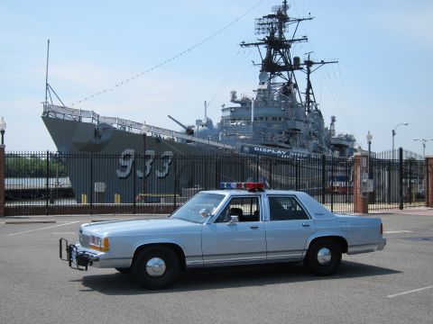 1991 Ford LTD Crown Victoria - Front-quarter view of a silver-blue police sedan with brush guard parked before a gray warship marked 933.