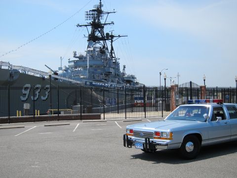 1991 Ford LTD Crown Victoria - silver-blue police sedan at right of frame parked before a moored gray Navy display warship marked 933.