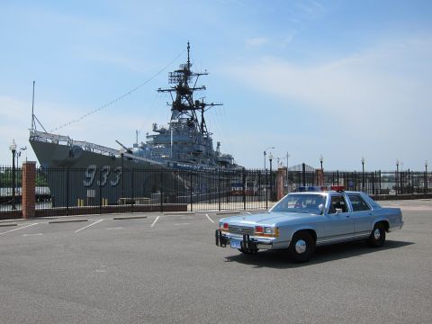 1991 Ford LTD Crown Victoria - silver-blue police sedan with push bar parked across an empty lot from a moored gray warship marked 933.