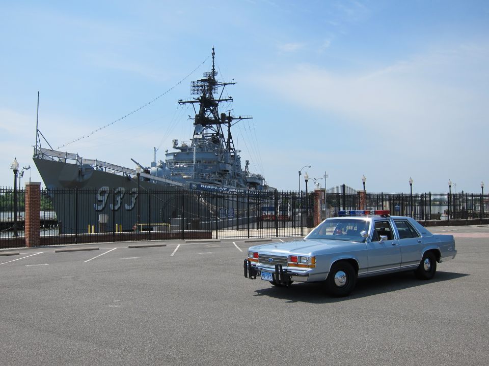 Silver-blue police sedan in lot before gray display warship 933