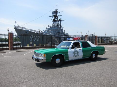 1991 Ford LTD Crown Victoria - green-and-white Sheriff sedan with star decal parked in a lot before a moored gray warship marked 933.