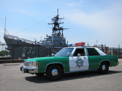 1991 Ford LTD Crown Victoria - Front-three-quarter view of a green-and-white Sheriff sedan parked before a moored gray warship marked 933.
