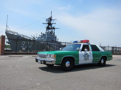 1991 Ford LTD Crown Victoria - green-and-white Sheriff sedan with roof lightbar parked in an open lot before a gray warship marked 933.