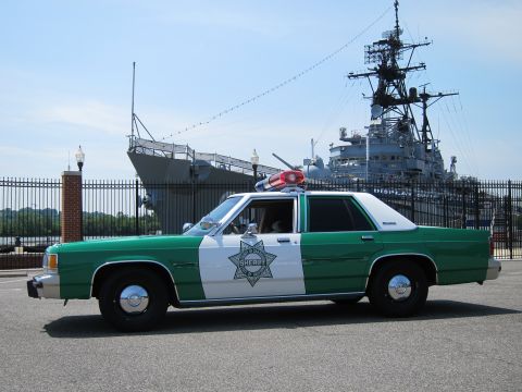 1991 Ford LTD Crown Victoria - Side profile of a green-and-white Sheriff sedan with red roof beacon parked before a moored gray warship.