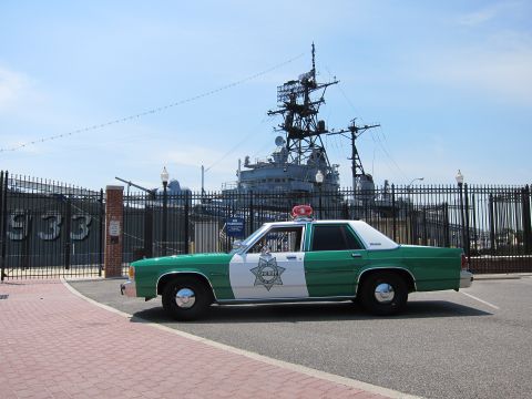 1991 Ford LTD Crown Victoria - green-and-white police sedan with a star door emblem sits on brick pavement before a gated gray naval warship marked 933.