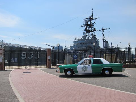 1991 Ford LTD Crown Victoria - green-and-white police sedan rounds a brick drive beside a black fence, with a museum warship numbered 933 looming behind.