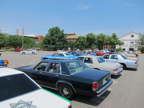 1991 Ford LTD Crown Victoria - Numerous vintage police sedans, including dark and two-tone units, fill a paved lot in front of brick and white buildings.