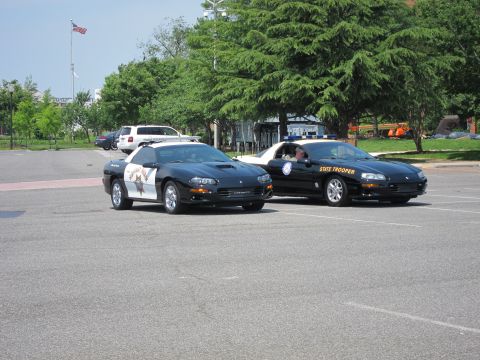 1991 Ford LTD Crown Victoria - Two black fourth-gen Chevrolet Camaro police pursuit cars, one lettered State Trooper, park nose-to-tail on a paved lot.