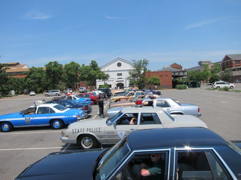 1991 Ford LTD Crown Victoria - crowded lot of classic police sedans, including a blue NYPD car and a Rhode Island State Police unit, before a white building.