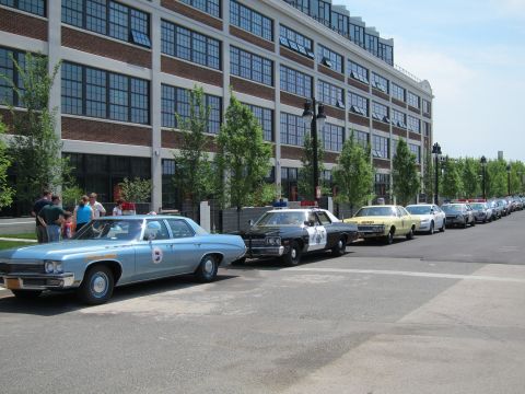 1991 Ford LTD Crown Victoria - long line of classic police cars, led by a light-blue sedan with a roundel, parked along a curb beside a multi-story brick building.